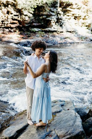 A couple embracing and laughing together while standing on rocks beside a rushing river in a natural setting.
