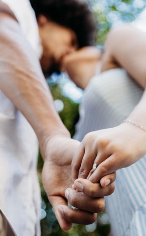 Couple sharing a tender kiss while holding hands outdoors, woman wearing an engagement ring and light blue dress.