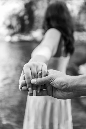 Black and white photo of couple holding hands near water, showcasing a diamond engagement ring.