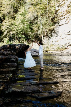 Bride and groom dancing on stepping stones in a forest creek, wearing white wedding attire.