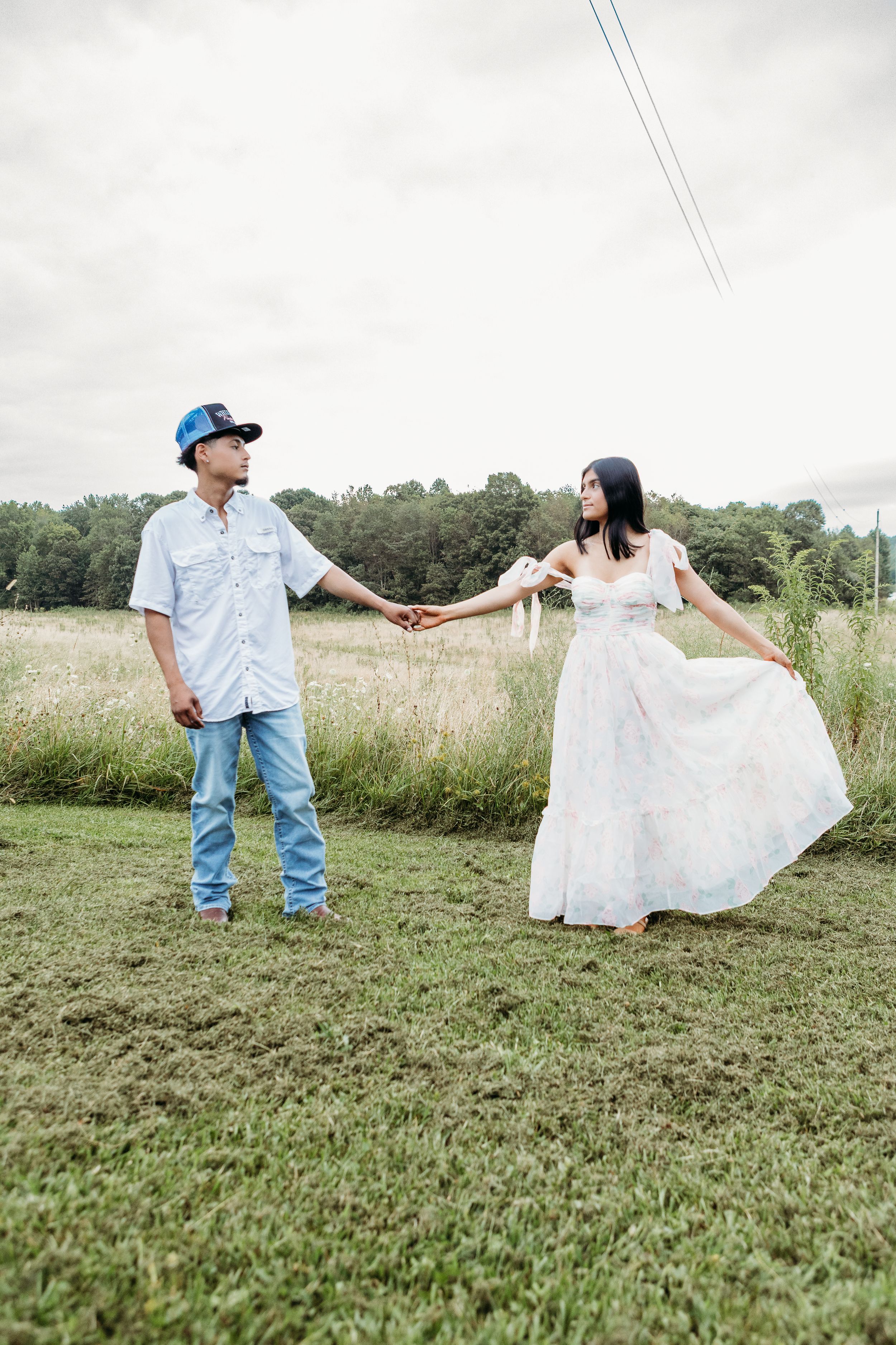 Engaged couple holds hands in a breezy field, woman in white floral dress, man in denim.