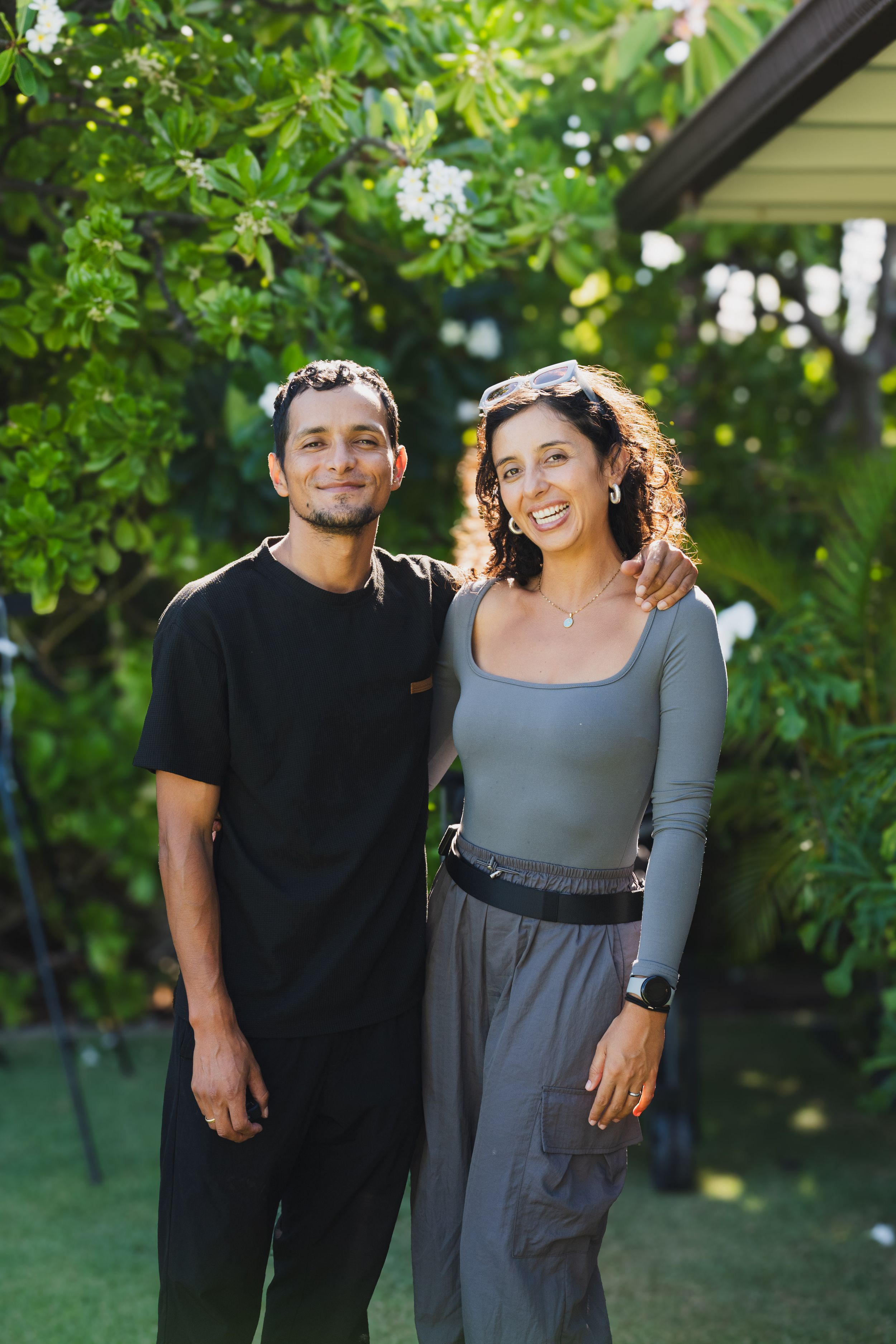 Two people stand together smiling in a garden setting with green foliage and a house in the background.