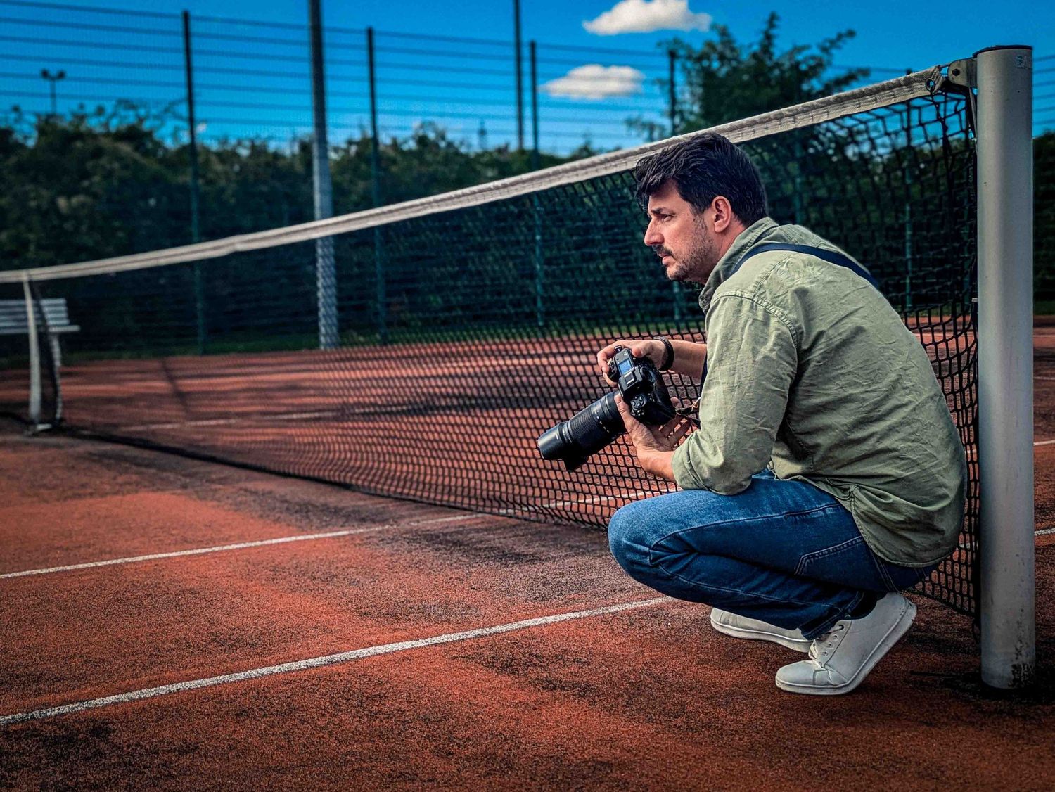 Photographer crouching at a tennis net on a clay court holding a professional camera.