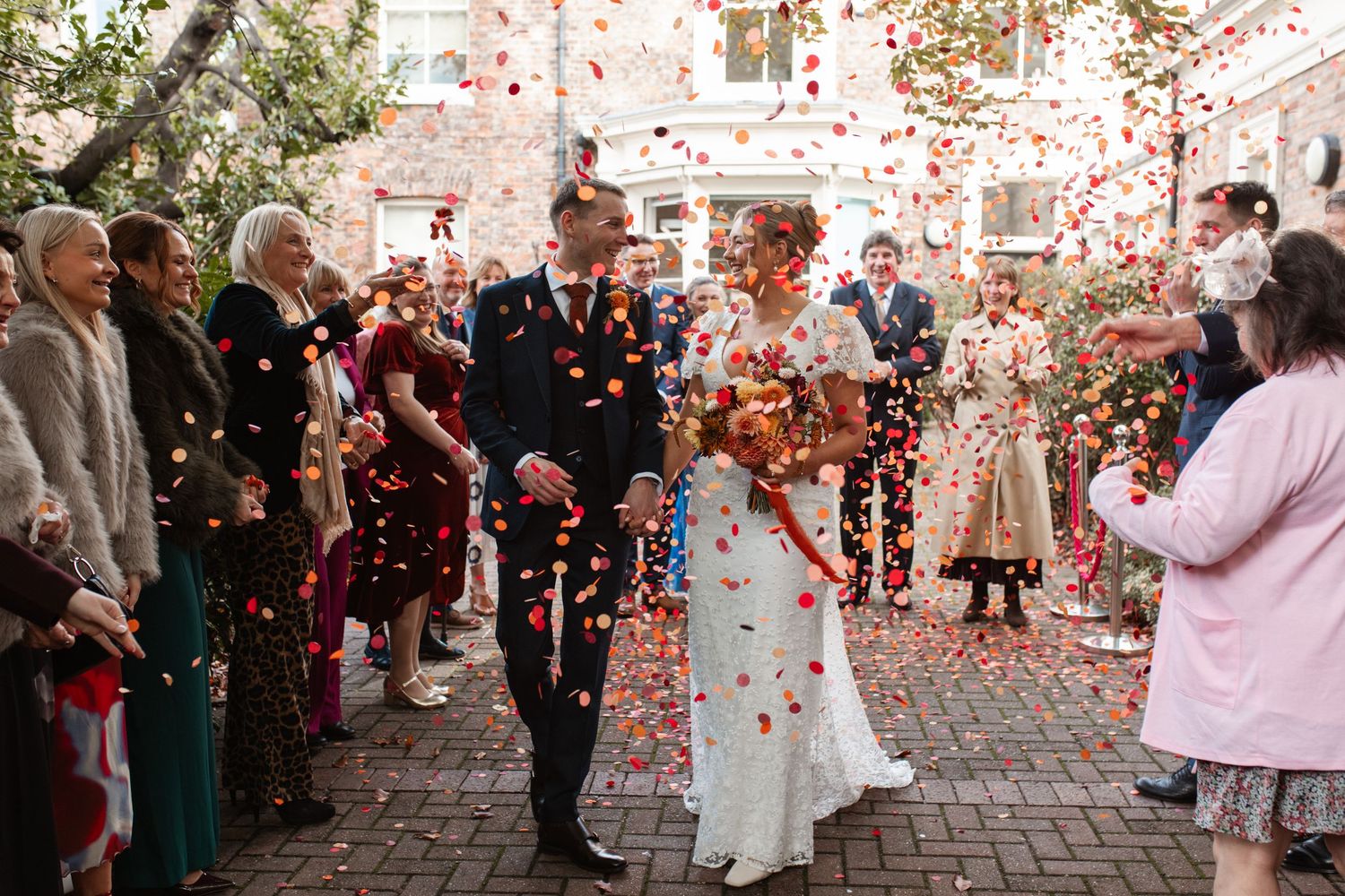 Red confetti fills the air during an outdoor celebration as people gather beneath trees and buildings.