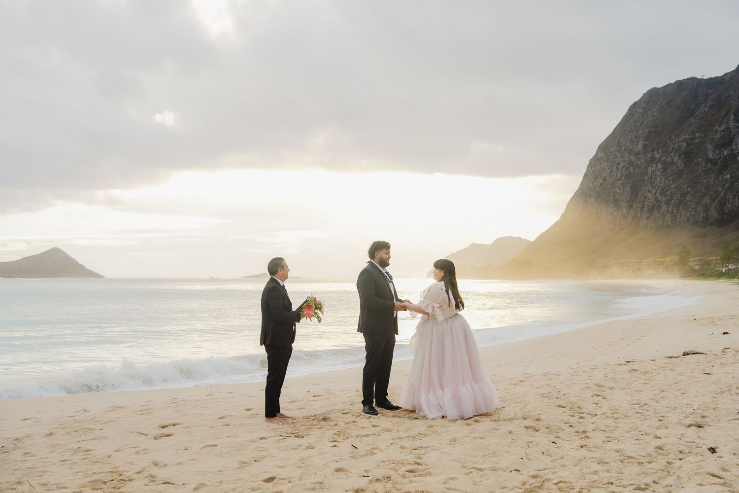 A couple in wedding attire walks along a misty beach at sunset with dramatic cliffs in the background.