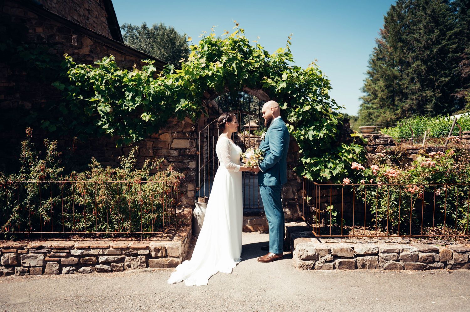 Couple de mariés posant devant un bâtiment en pierre recouvert de vigne lors de leur mariage en extérieur.