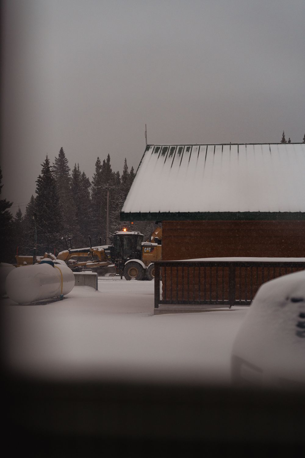 Yellow CAT motor grader clearing snow near snow-covered log cabin building with pine trees behind.