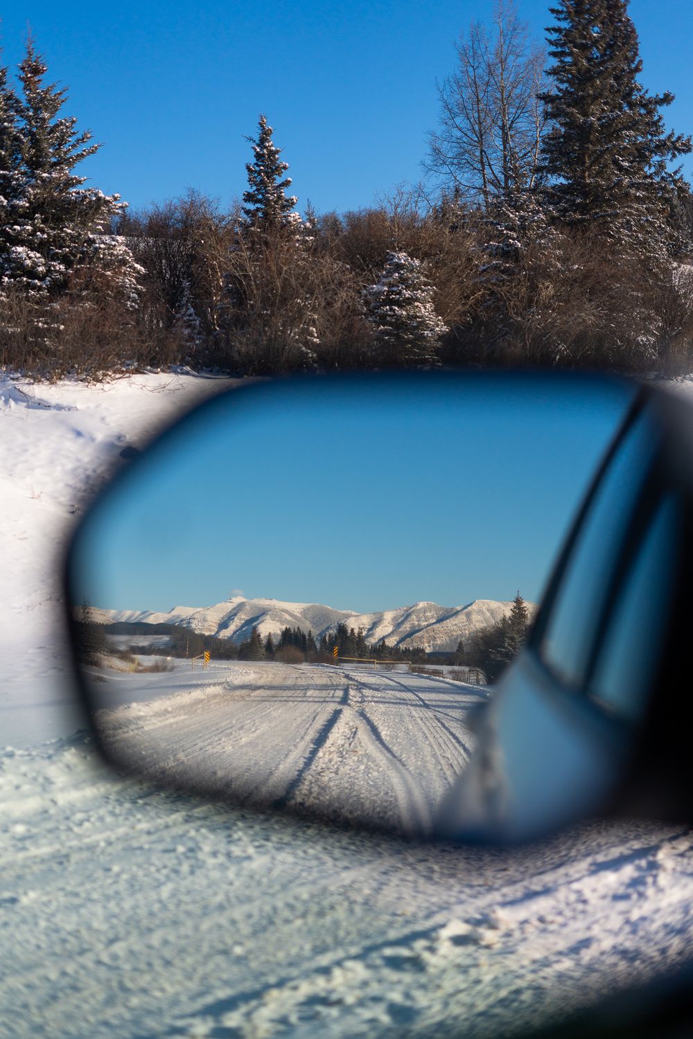 Snowy winter road reflected in a car side mirror with mountains and evergreen trees under a clear blue sky.