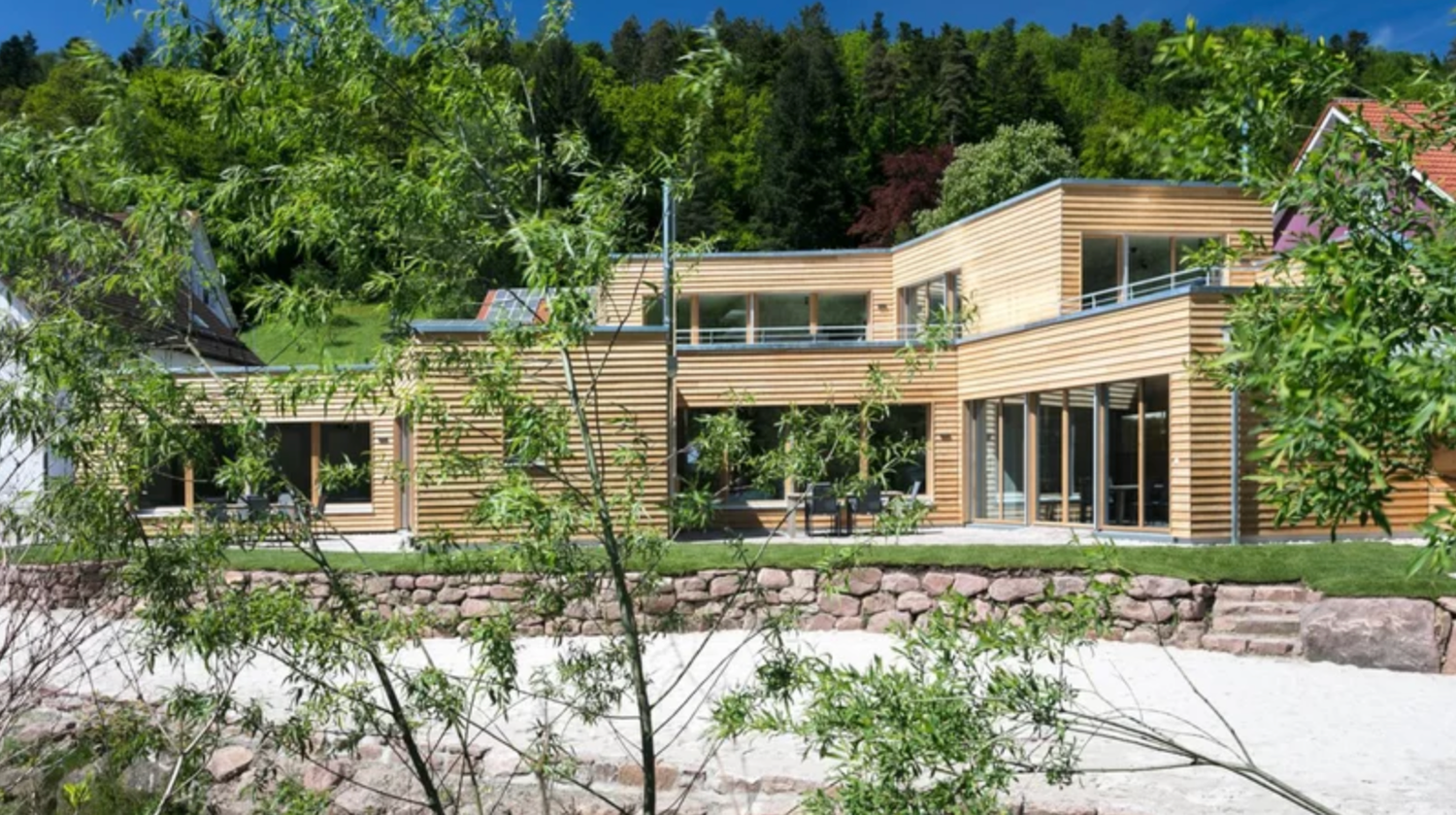 A panoramic view of a modern wooden home nestled in lush greenery with large windows and stone landscaping.