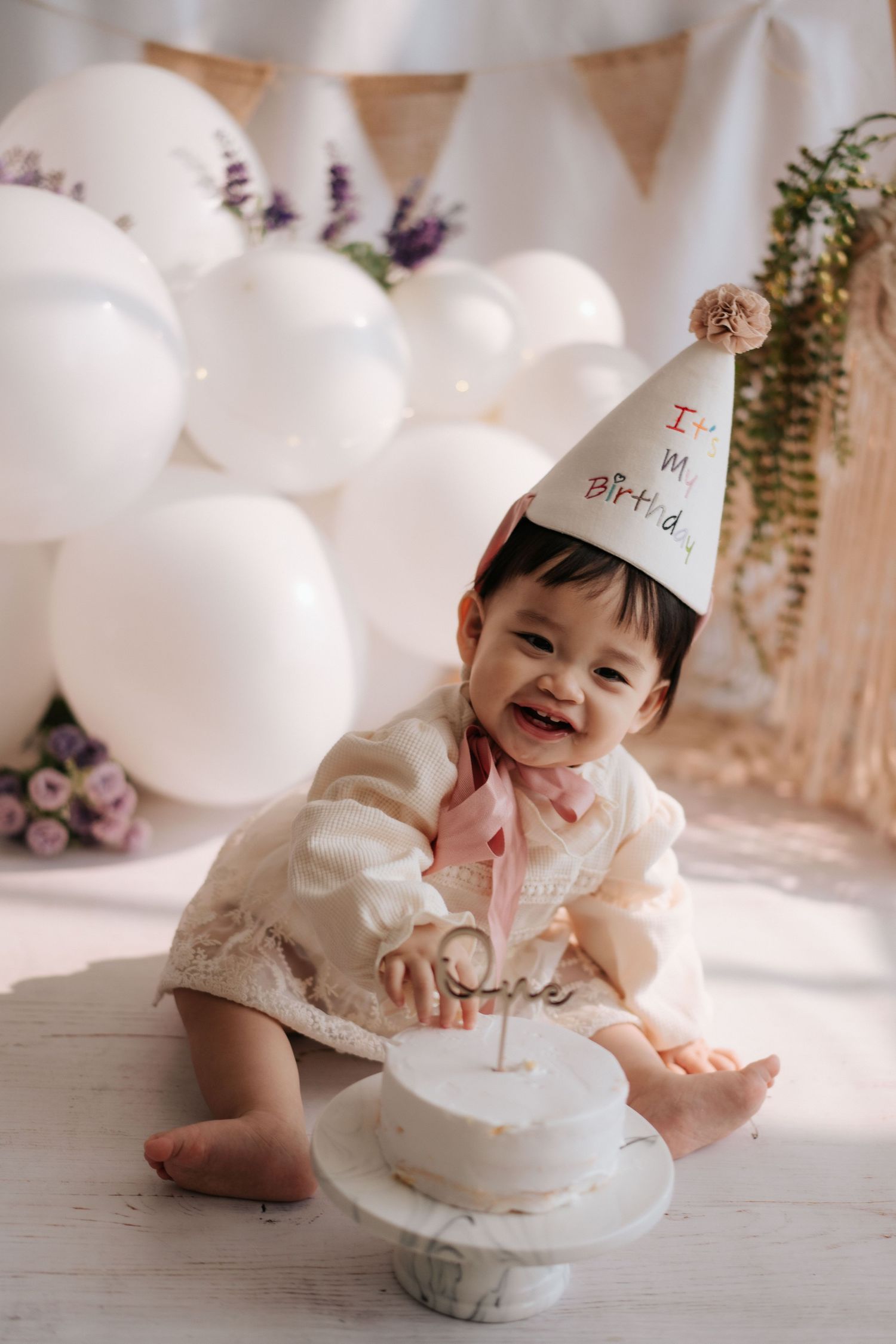 Baby in pink dress celebrates first birthday with smash cake surrounded by greenery and party decorations.