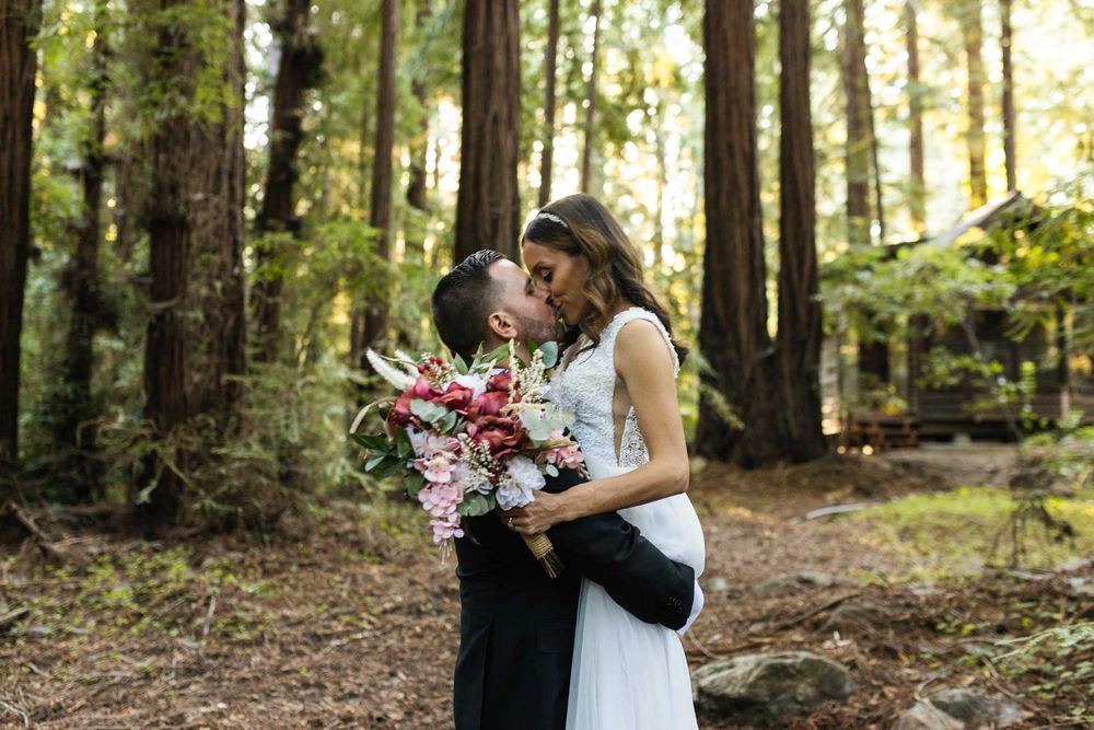 Wedding couple shares an intimate moment among tall trees in a sunlit forest setting.