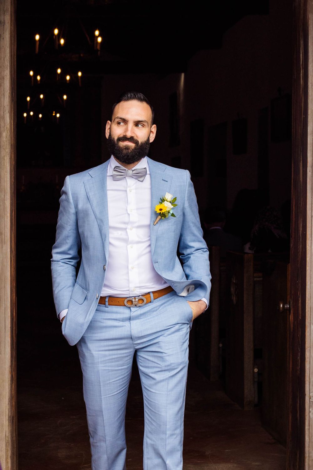 Groomsmen in light blue suits with yellow boutonnieres pose in a rustic venue with string lights.