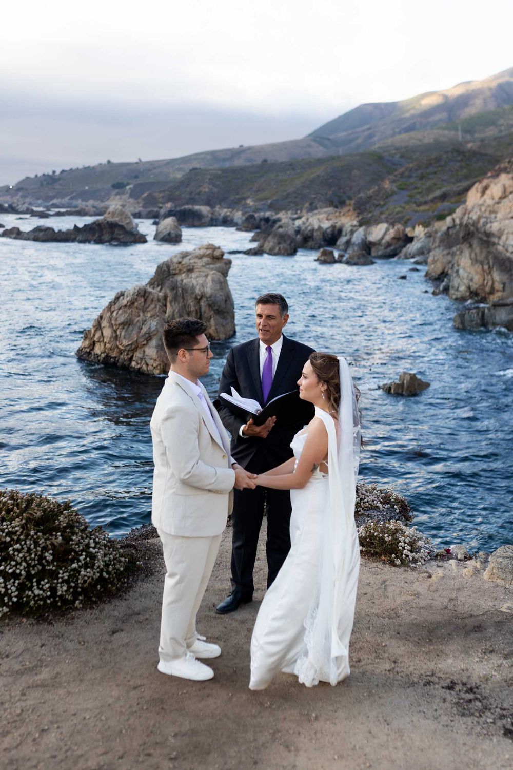 A wedding ceremony takes place on a dramatic coastal cliff overlooking the ocean and rocky shoreline.