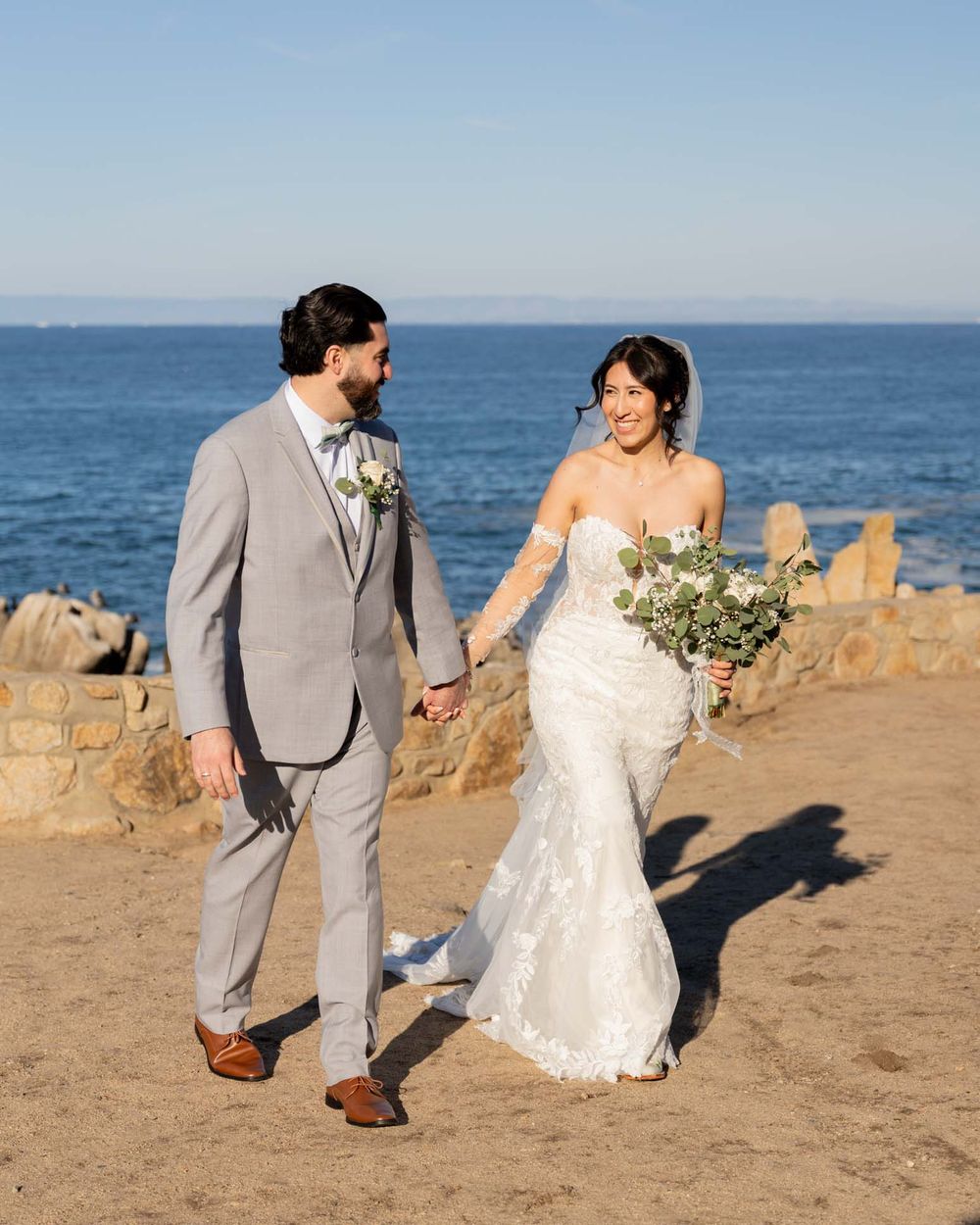Couple walking along sandy coastal path with ocean views wearing wedding attire on their special day.