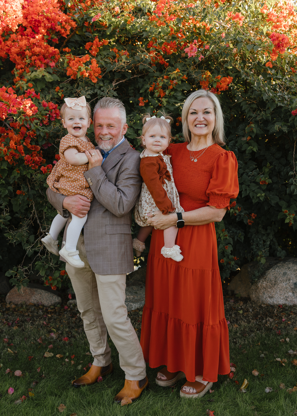 Grandparents holding two baby girls in autumn garden with red flowers.