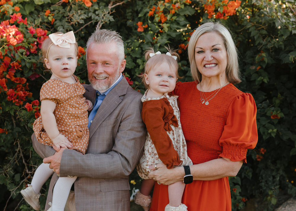 Smiling couple holding two toddler girls outdoors near red flowering bush.