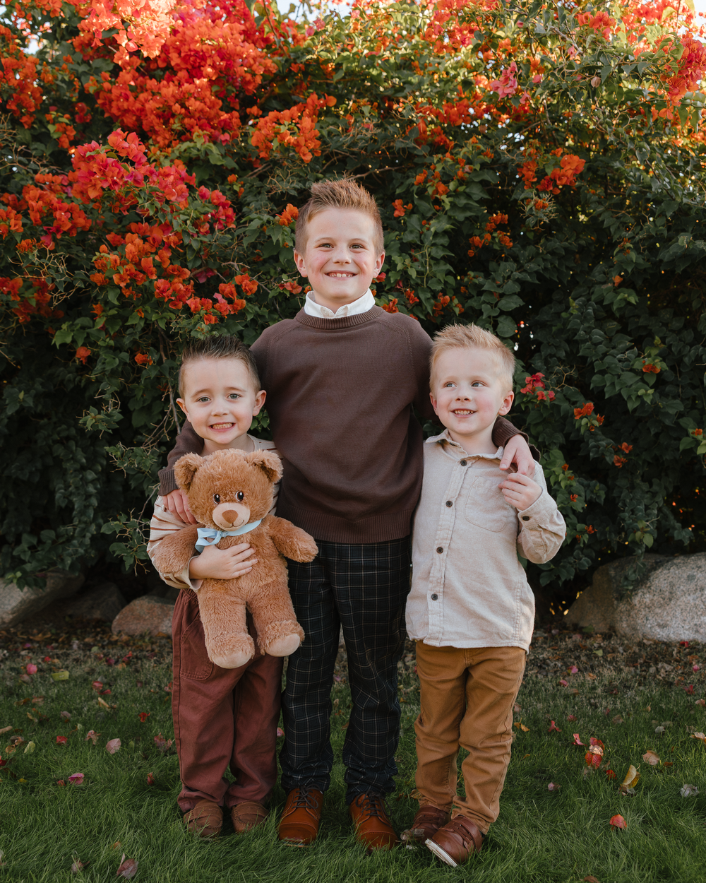 Three young brothers hugging and smiling in front of red flowering bush.