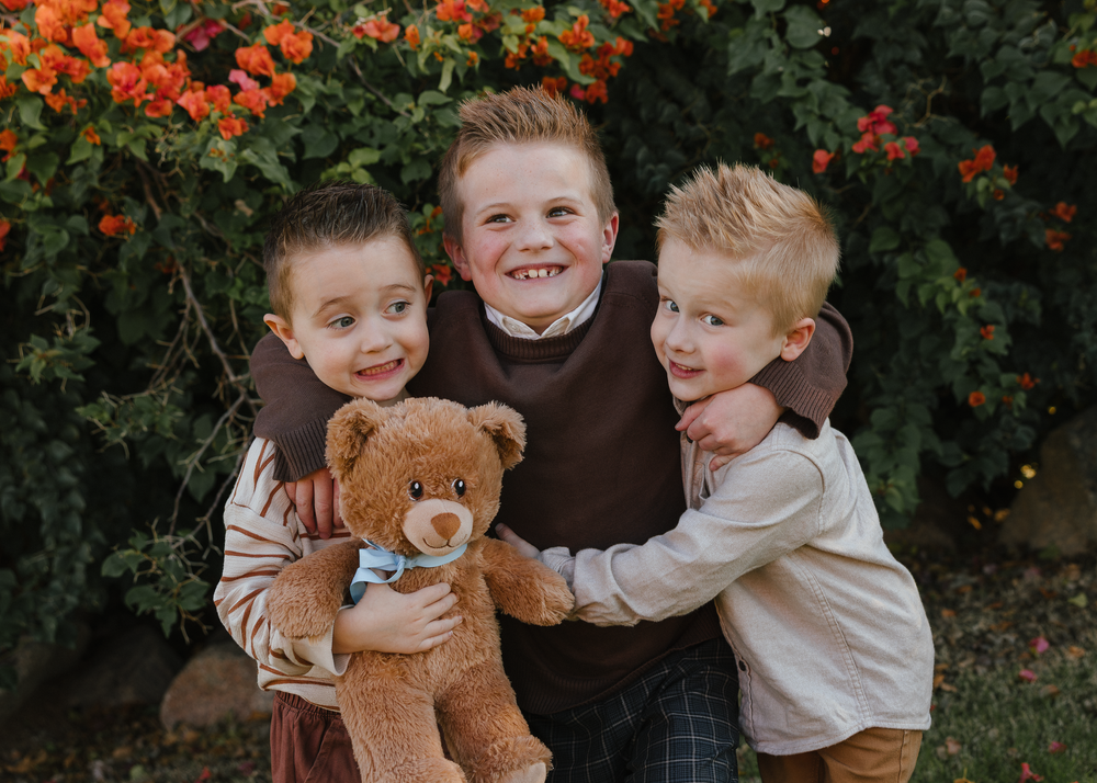 Three boys laughing and embracing with teddy bear near colorful shrubs.
