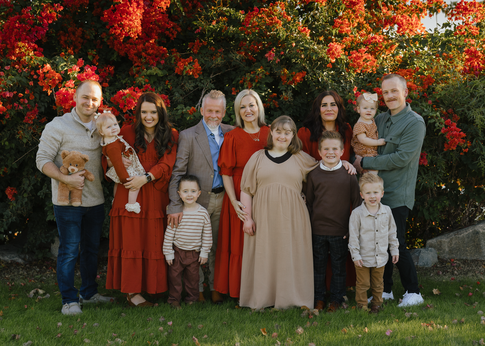 Extended family of ten posing together in front of vibrant red flowering bushes outdoors.