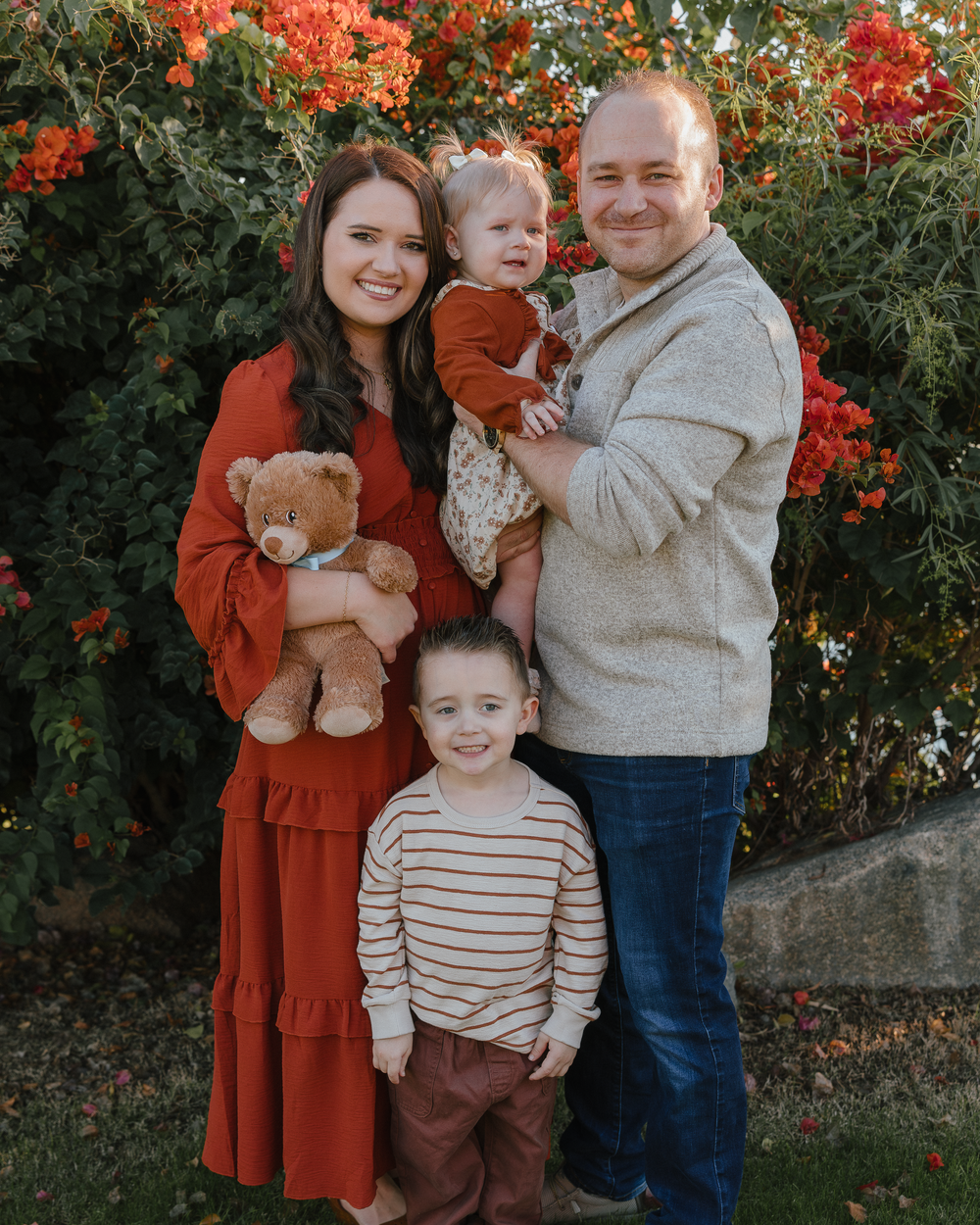 Young family of four posing with teddy bear in front of red blooms.