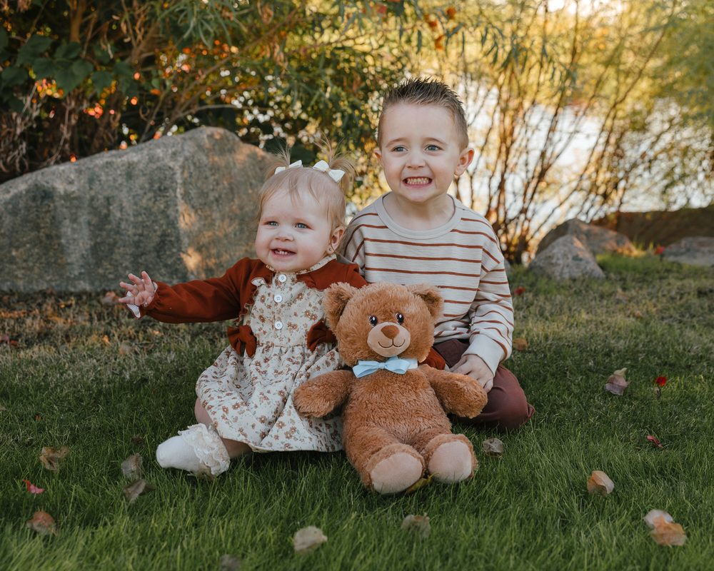 Two young siblings sitting on grass with teddy bear in autumn garden.
