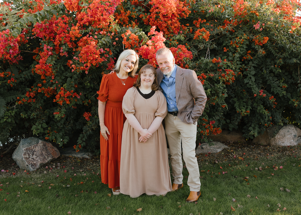 Two women and a man smiling together in front of red flowering bush.
