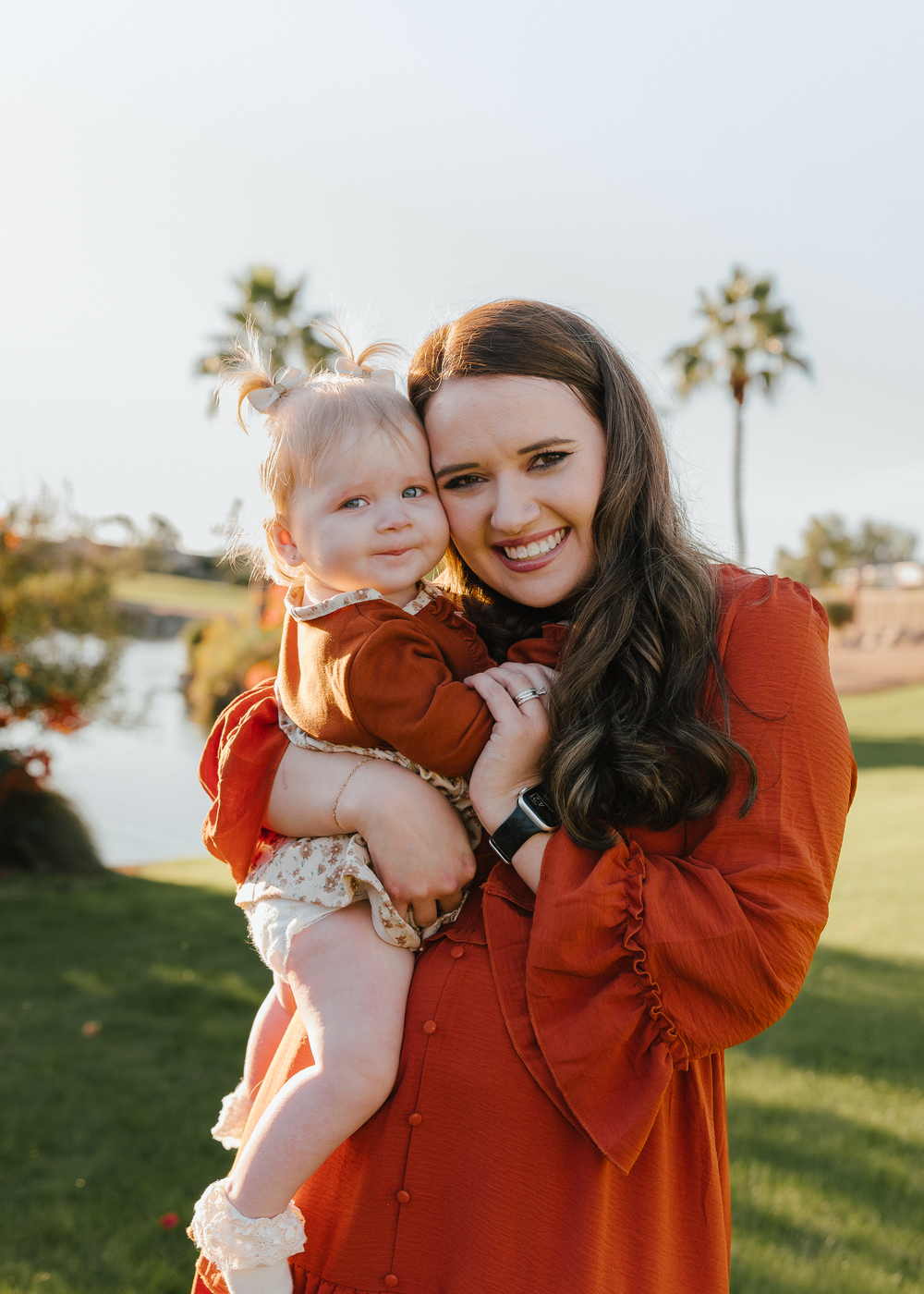 A smiling woman in a rust orange dress holds a baby girl outdoors near a pond with palm trees.