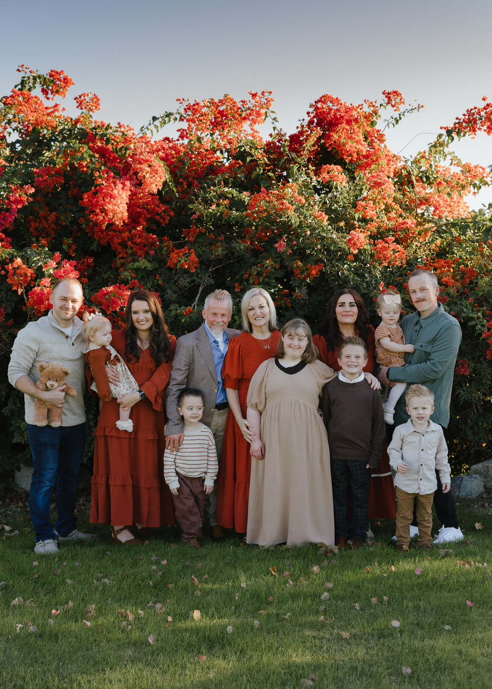 Large family portrait in front of vibrant red flowering bush outdoors.