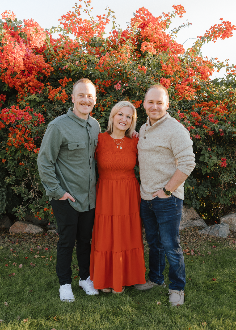 Three adults pose smiling in front of vibrant red flowering bushes in a garden setting.