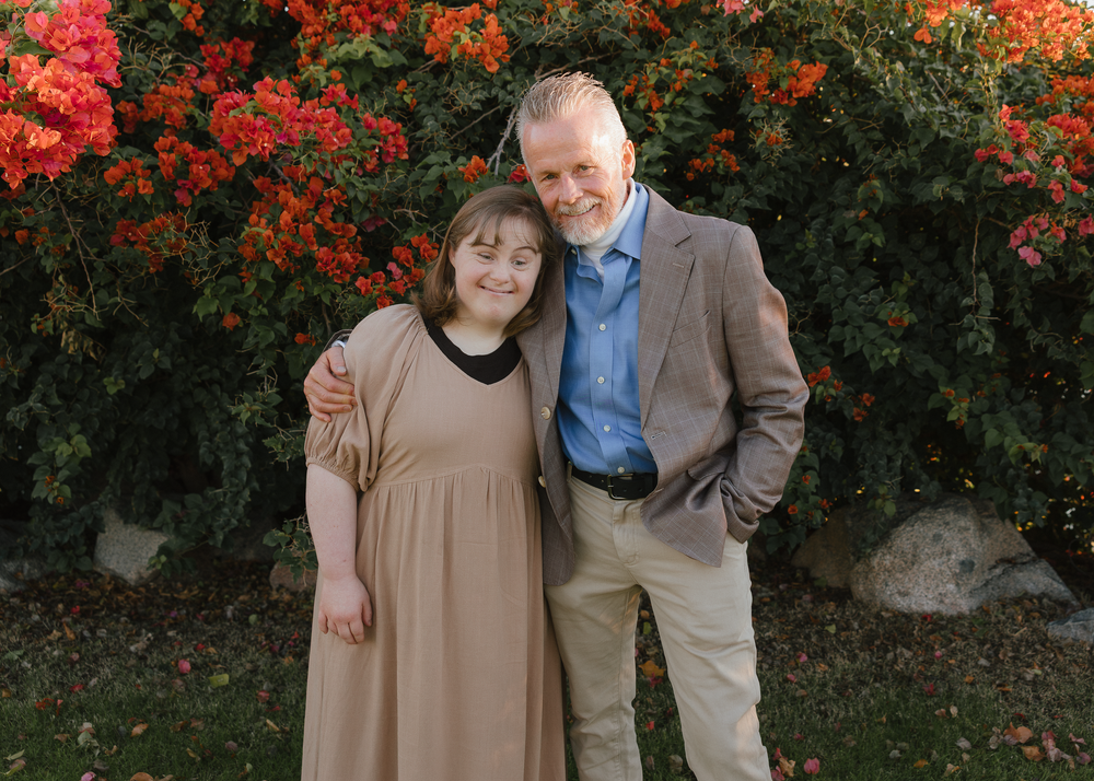 A young woman and older man with gray hair smile together in front of red flowering bushes.