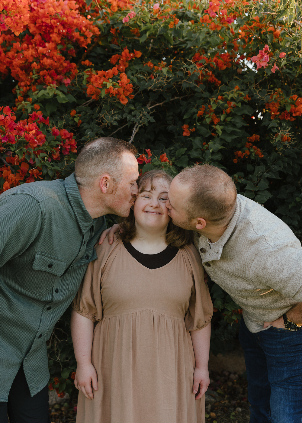 Two men kiss the cheeks of a smiling young woman in a tan dress in front of red flowers.