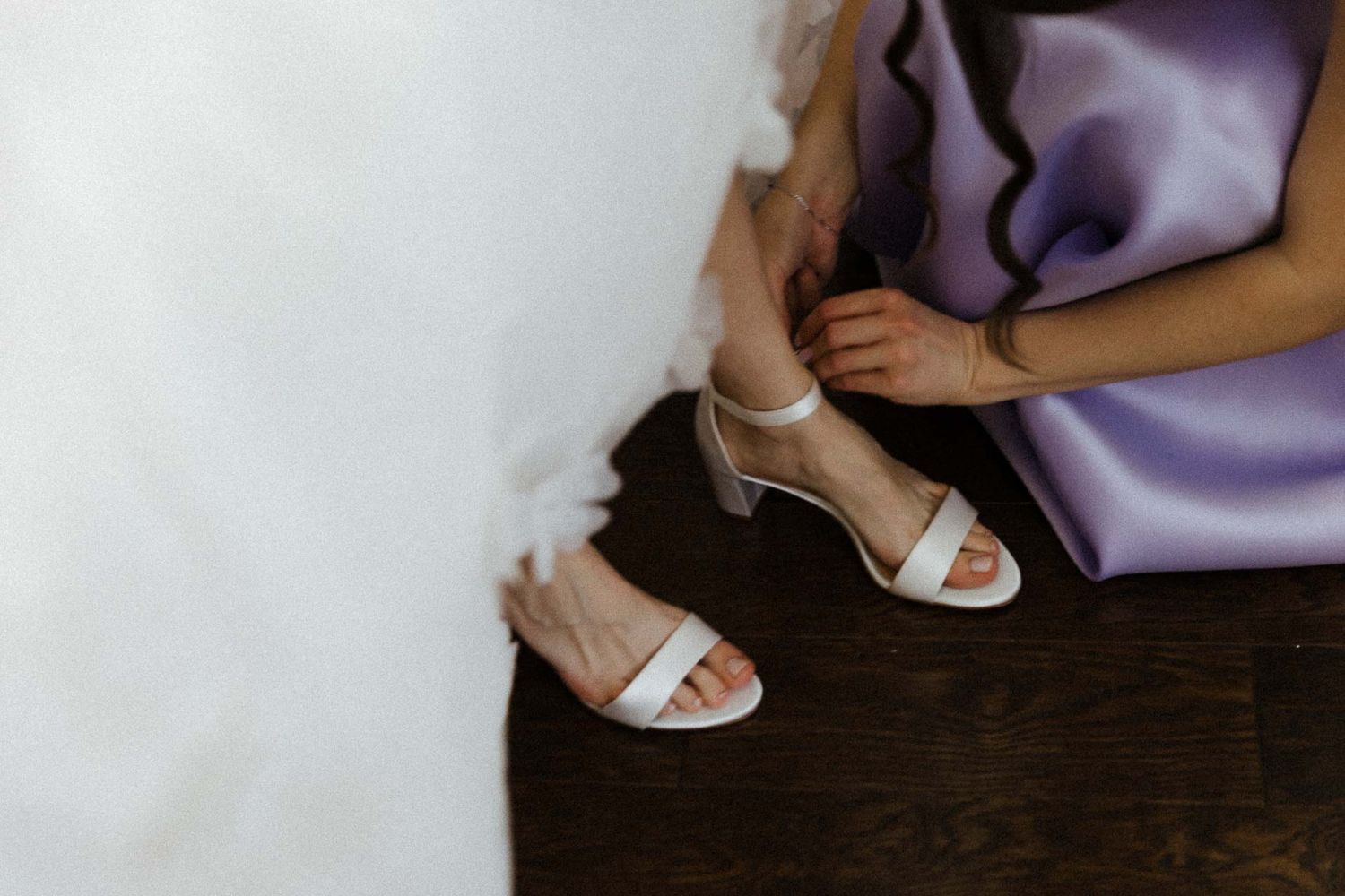 White bridal sandals on a dark wooden floor next to flowing purple fabric.
