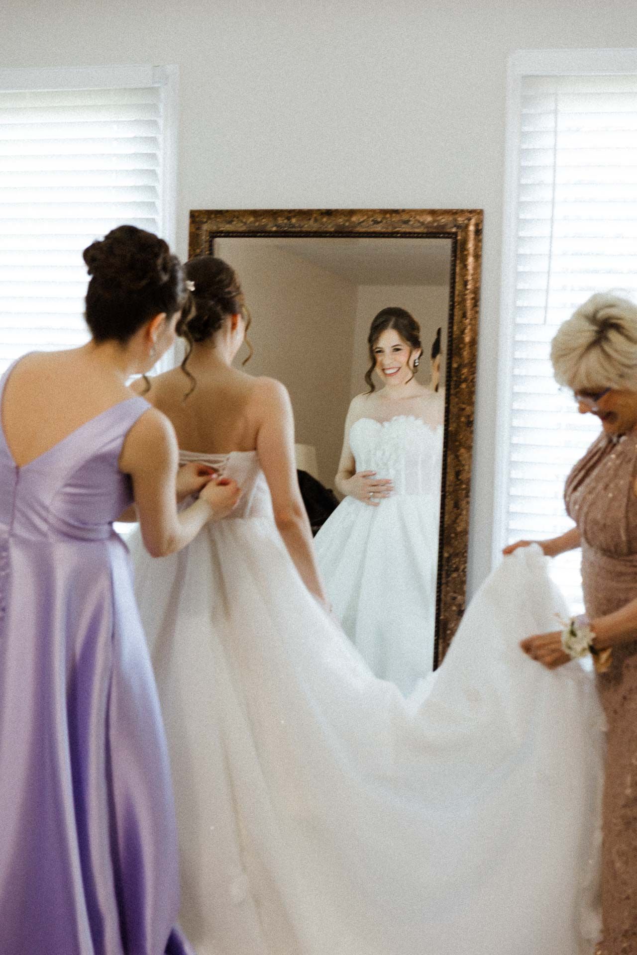 A bridal party helps with wedding dress preparations while looking in an ornate gold-framed mirror.