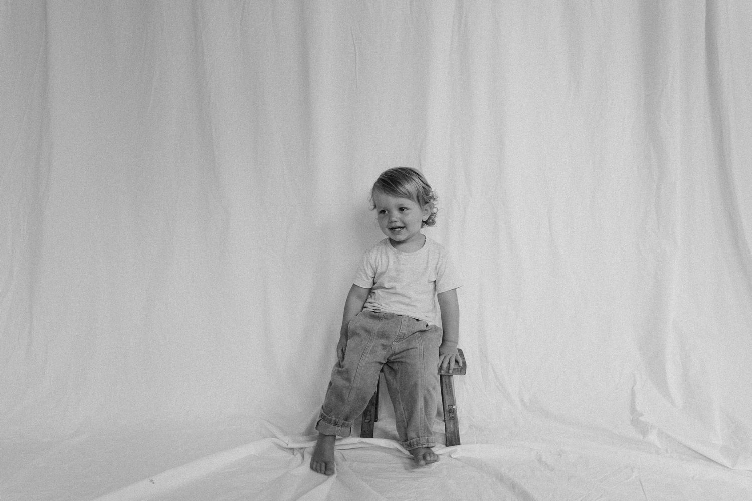 Black and white photo of smiling toddler in white shirt standing on white fabric backdrop in rustic studio in New Zealand.