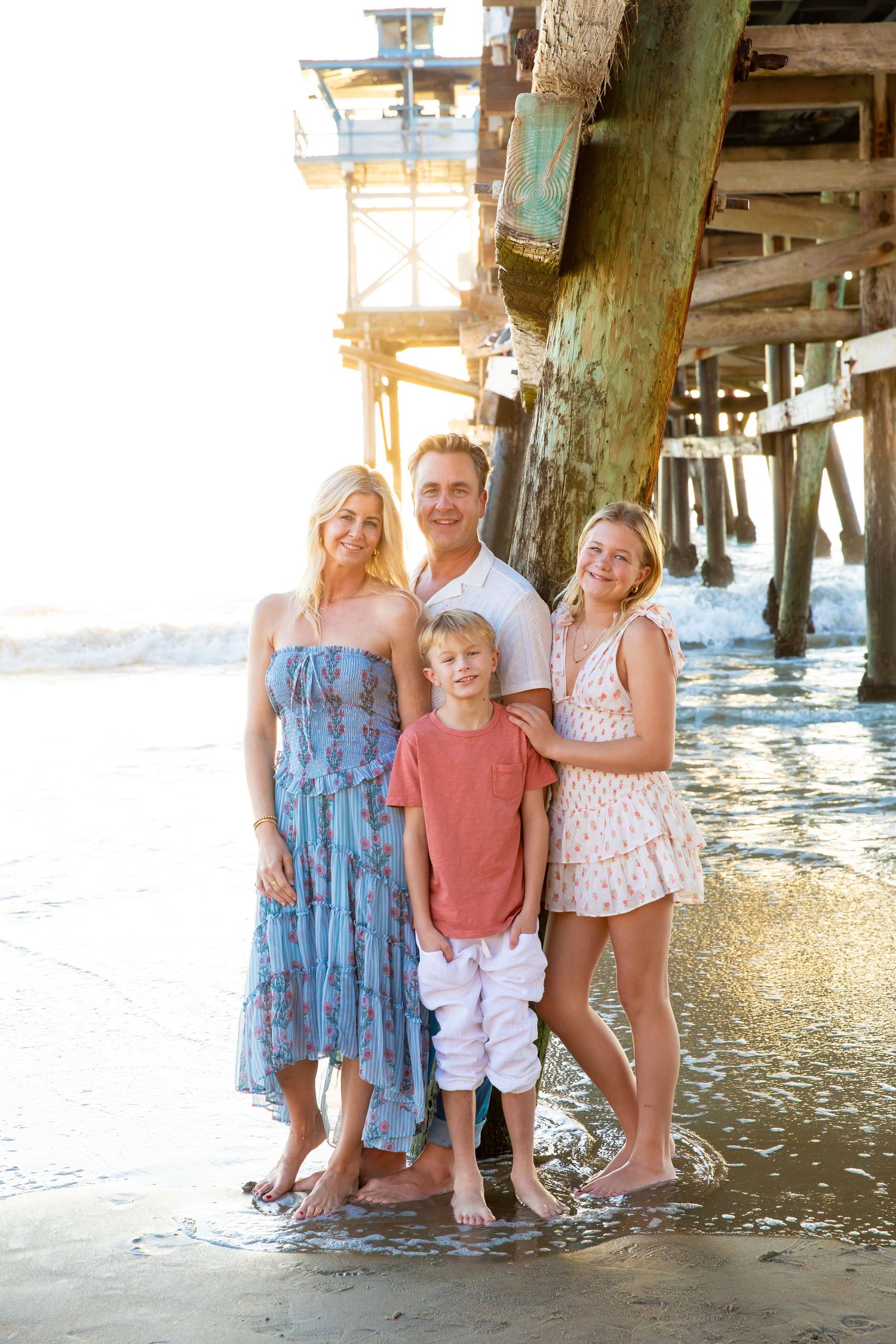 A family poses together under a wooden pier at the beach during sunset, wearing casual summer attire.