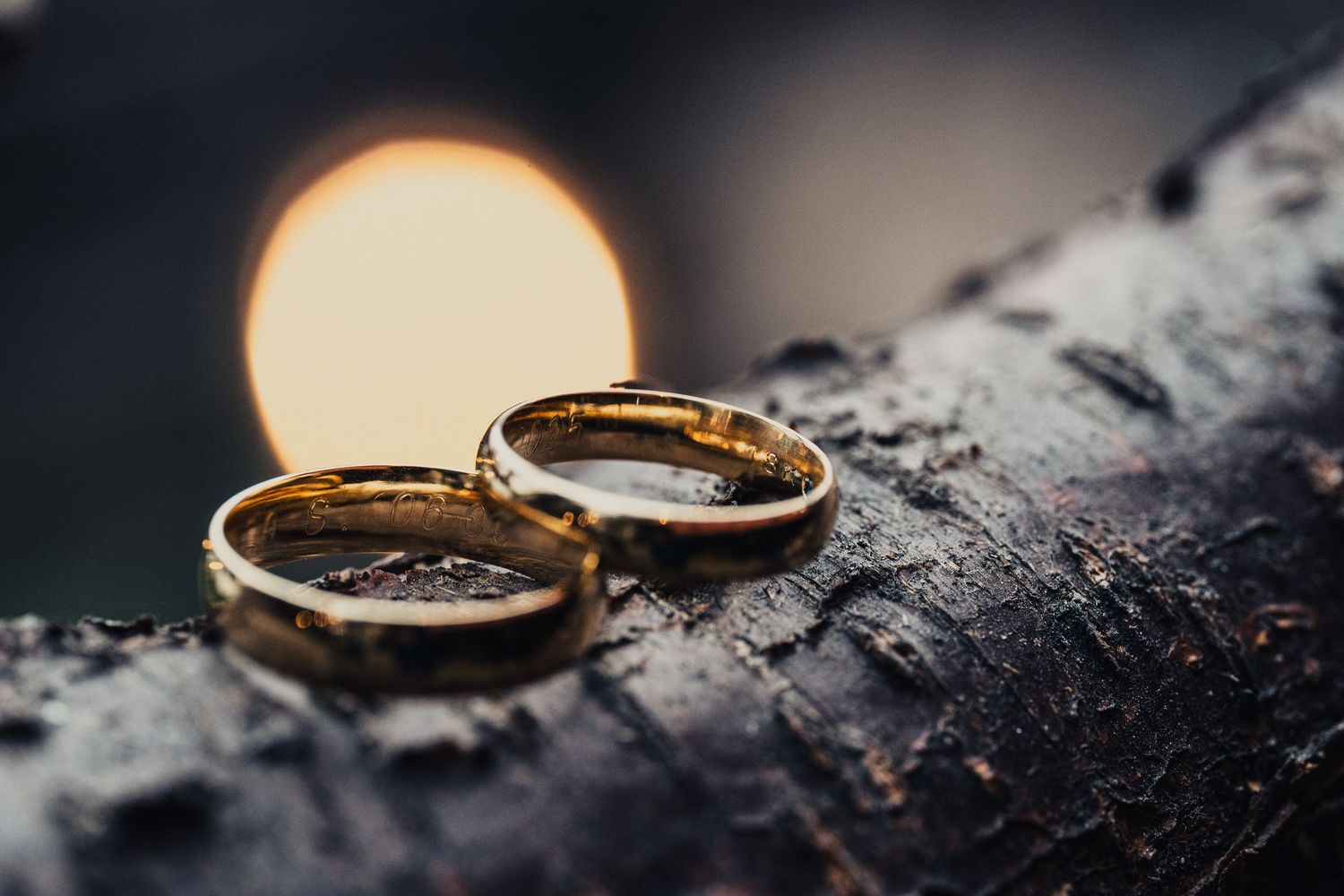A gold wedding ring rests on a dark textured surface with dramatic lighting creating a warm glow.