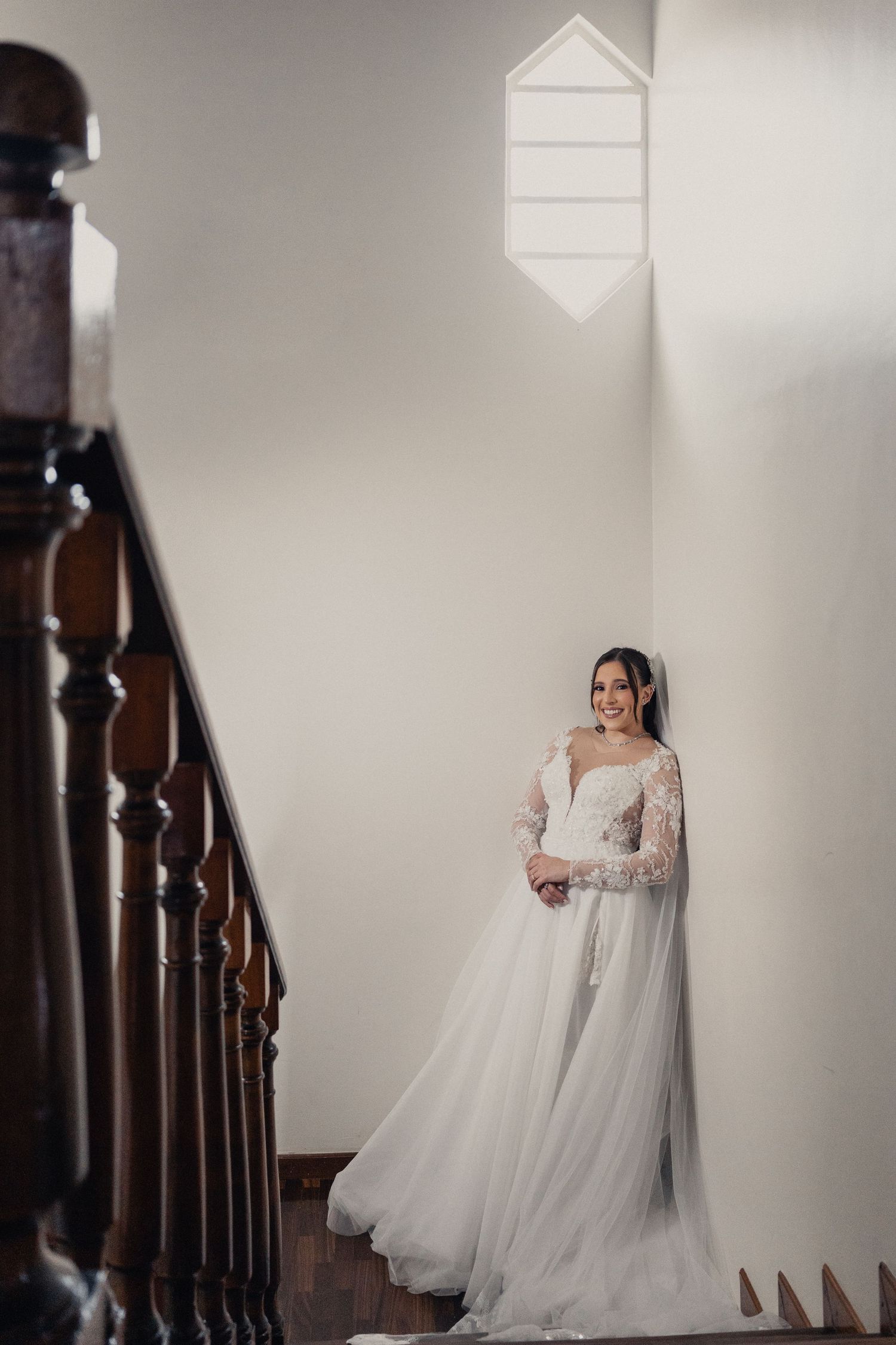 A bride in a flowing white wedding gown stands elegantly on a wooden staircase in soft indoor lighting.