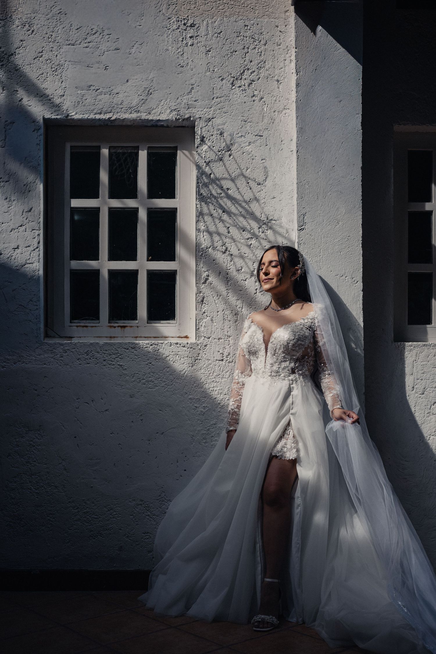 A bride in a lace wedding dress poses against a white wall with dramatic shadows and window light.