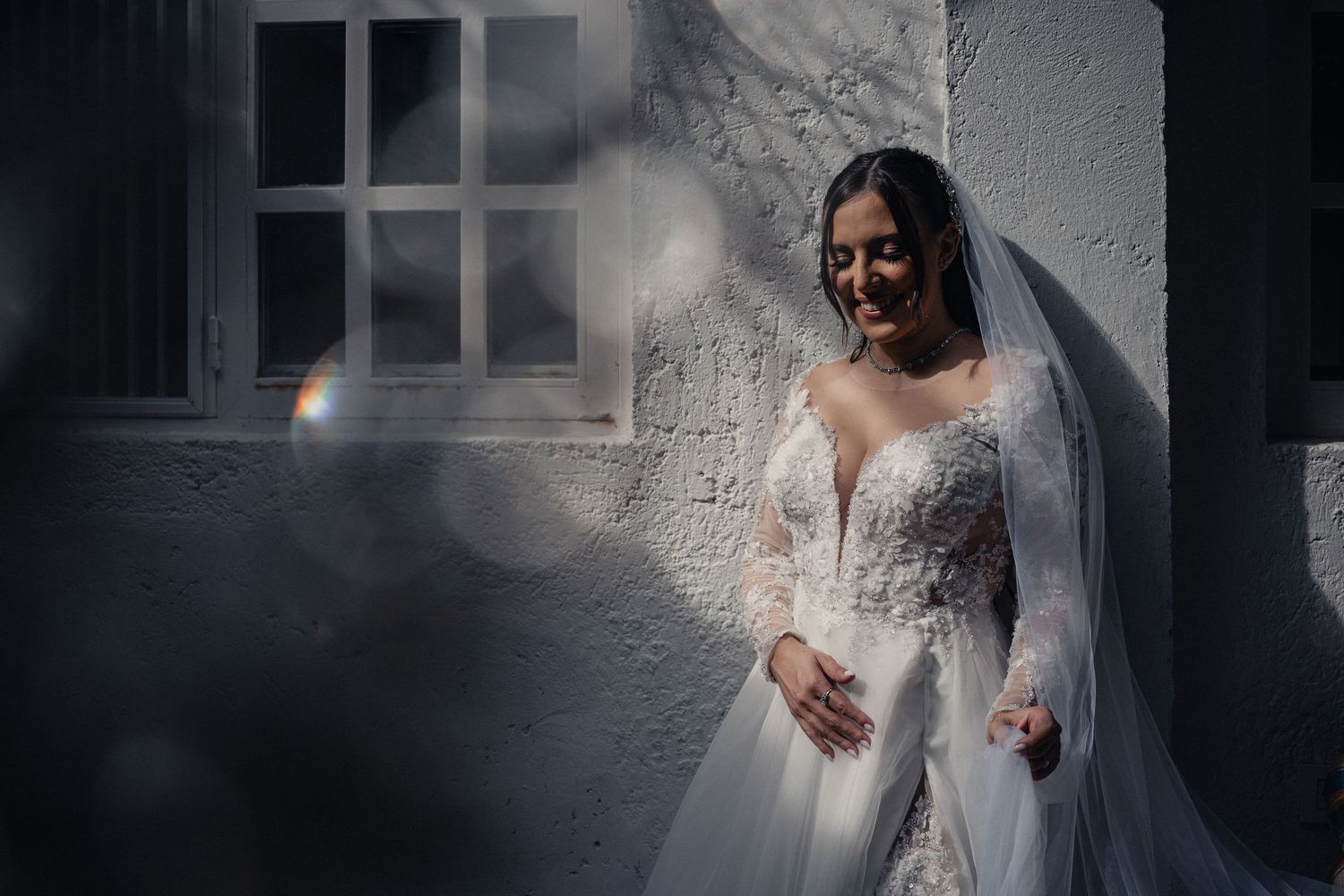 A bride in a white lace wedding dress and veil stands against a textured wall in dramatic lighting.