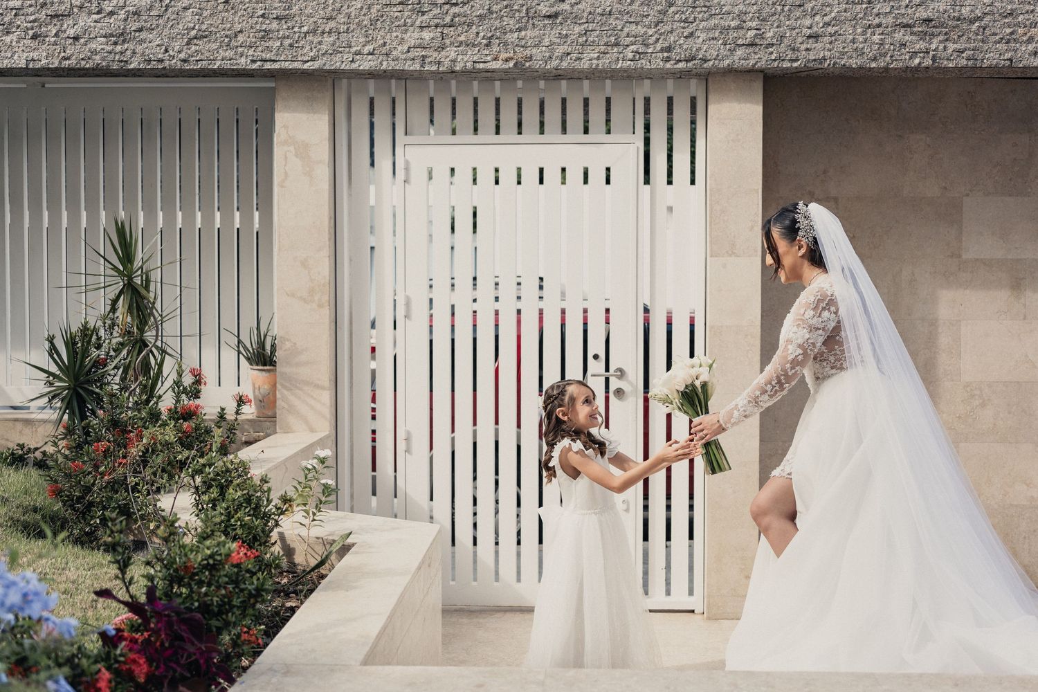 A small figure in a white dress shares a sweet moment with someone in an elegant white wedding gown on a porch.