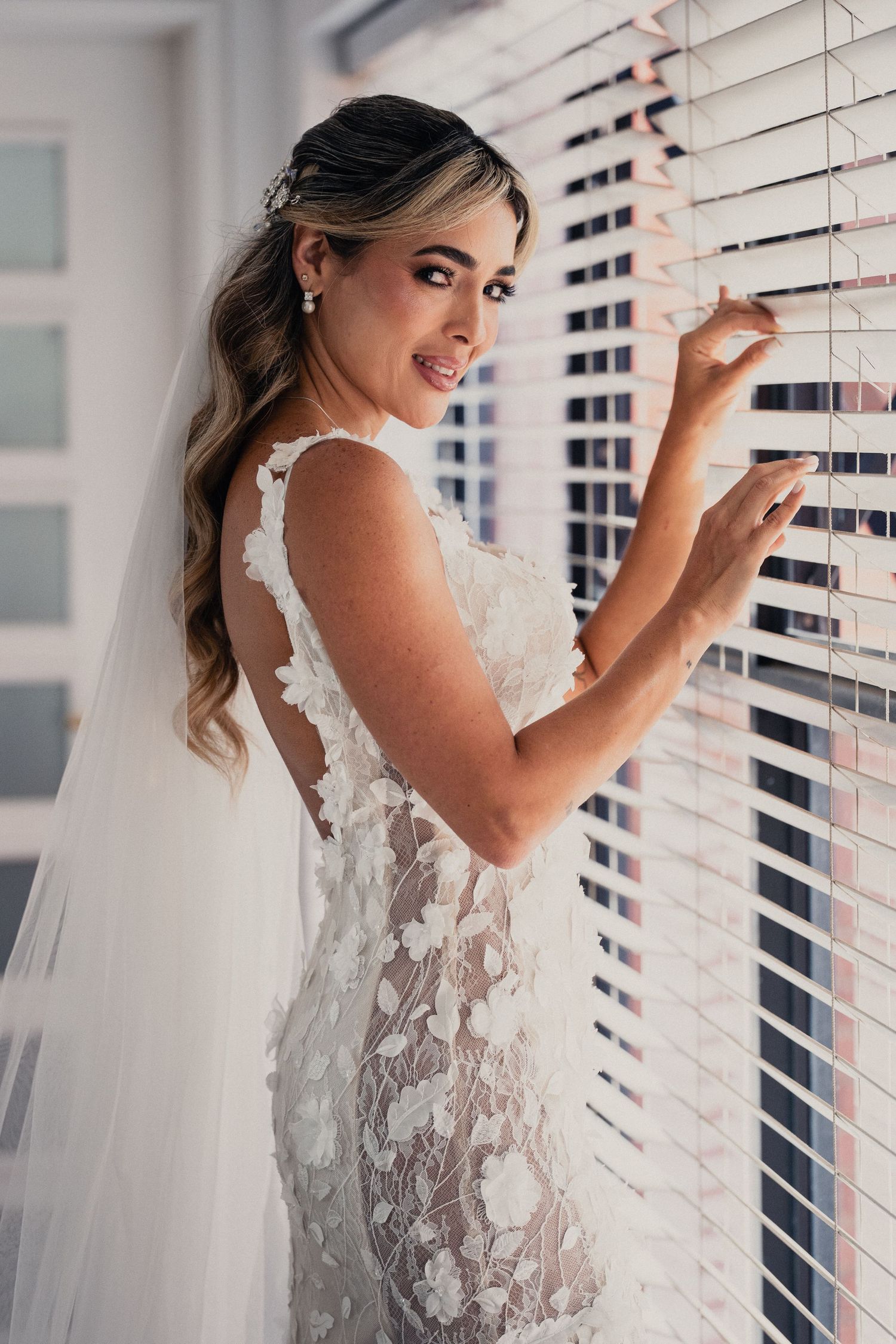 Bride in lace wedding dress poses playfully by window blinds in natural light.
