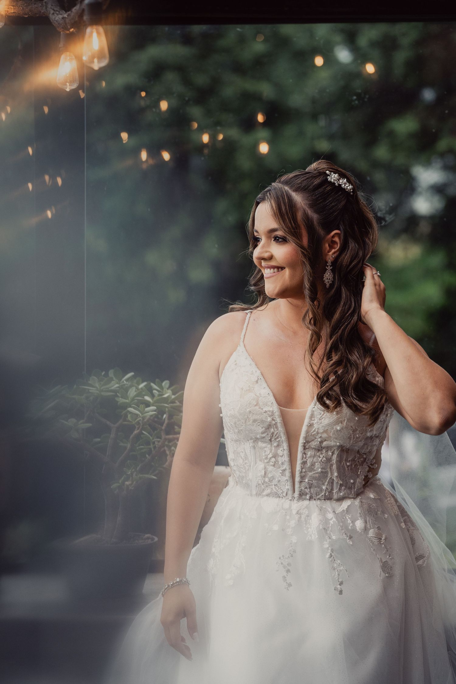 Bride in white lace wedding dress with plunging neckline poses outdoors with twinkling lights and smoke effects.