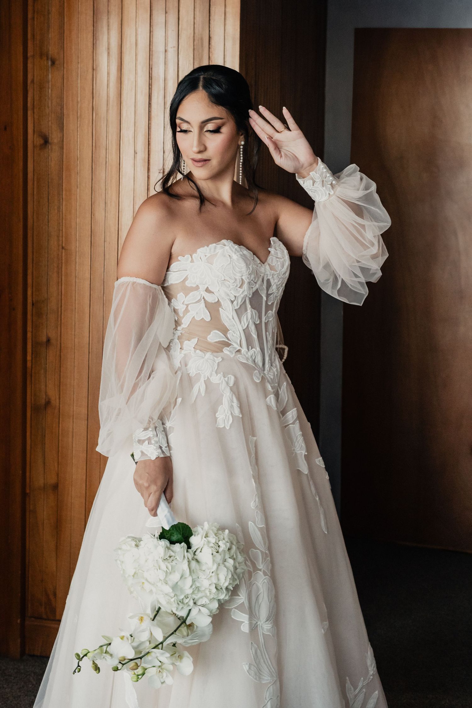 Elegant white wedding dress with delicate floral appliques and sheer off-shoulder sleeves in a wooden room.