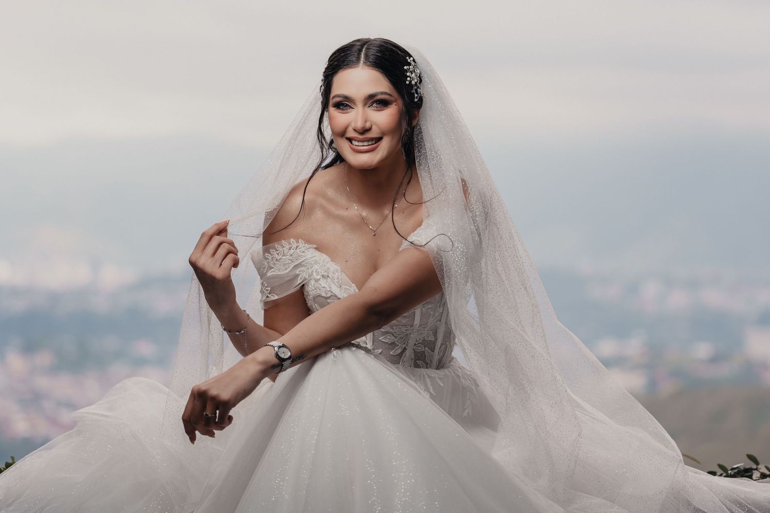 A bride in a flowing white wedding dress stands against a blurred mountain landscape backdrop.