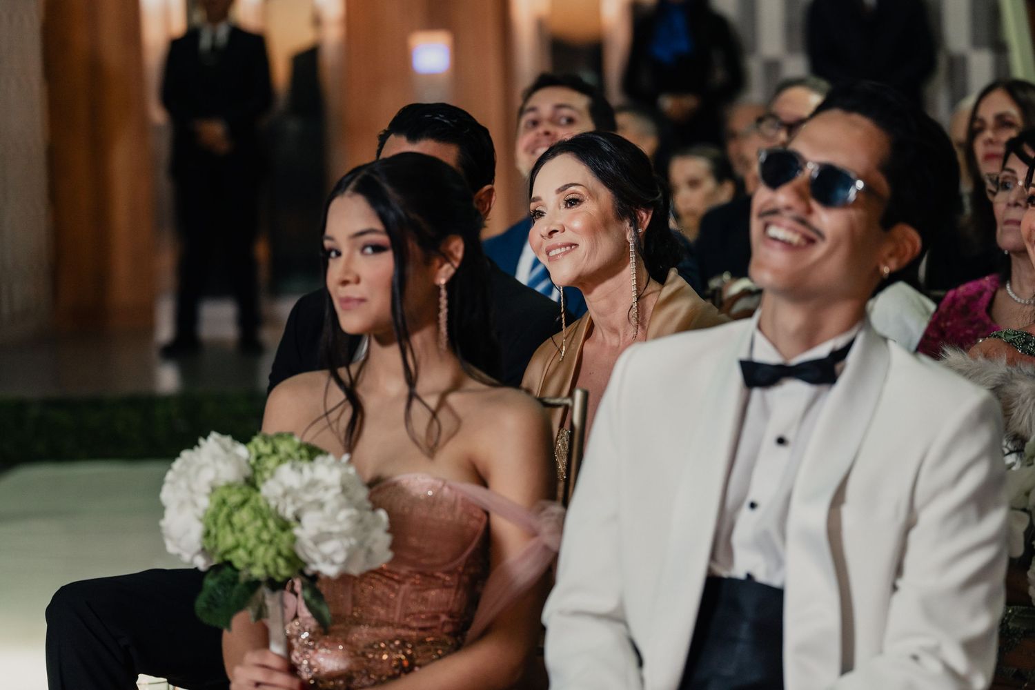 Wedding guests seated at a ceremony share joyful expressions while watching the proceedings.
