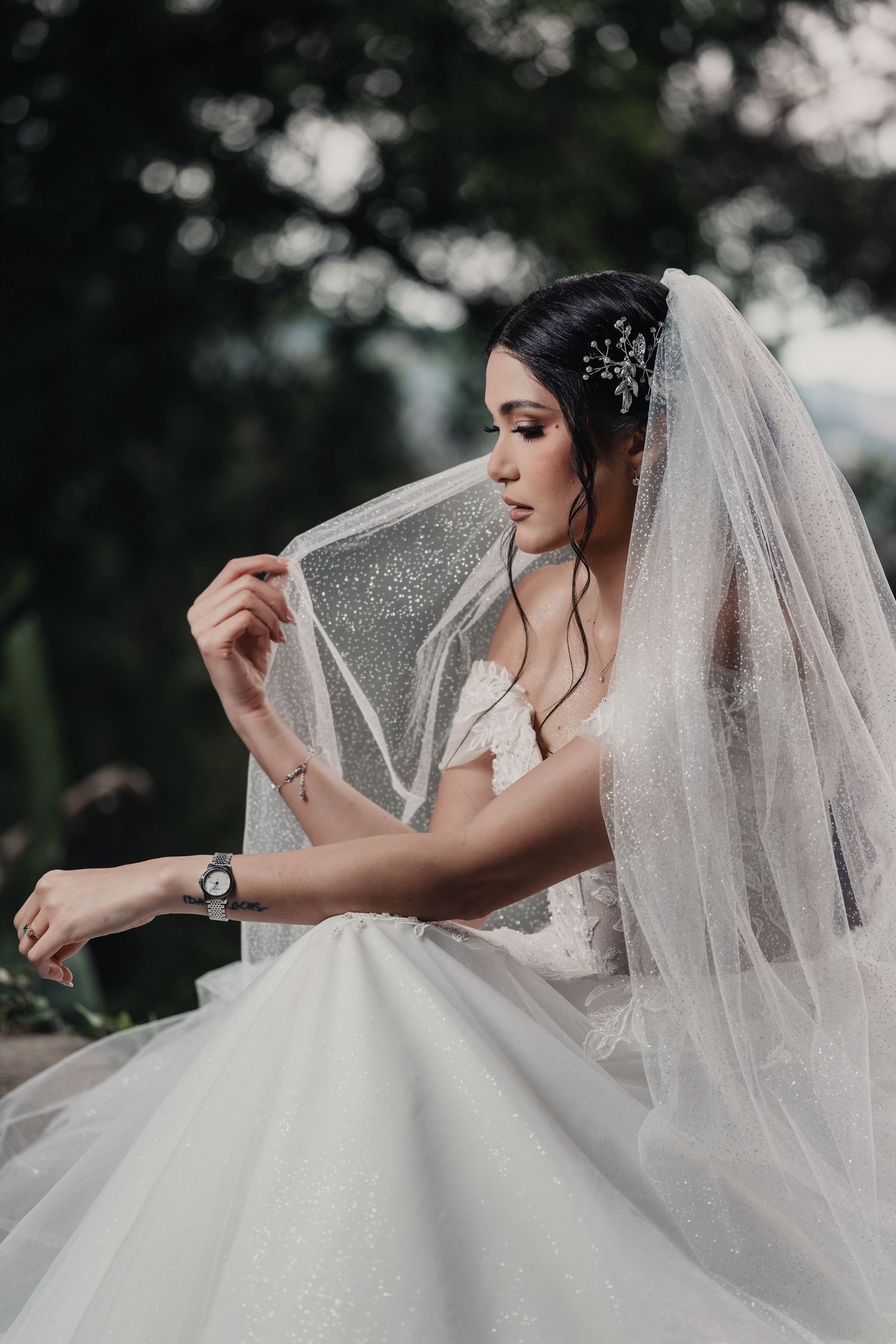 A bride in a flowing white wedding dress poses elegantly with her veil in an outdoor setting.