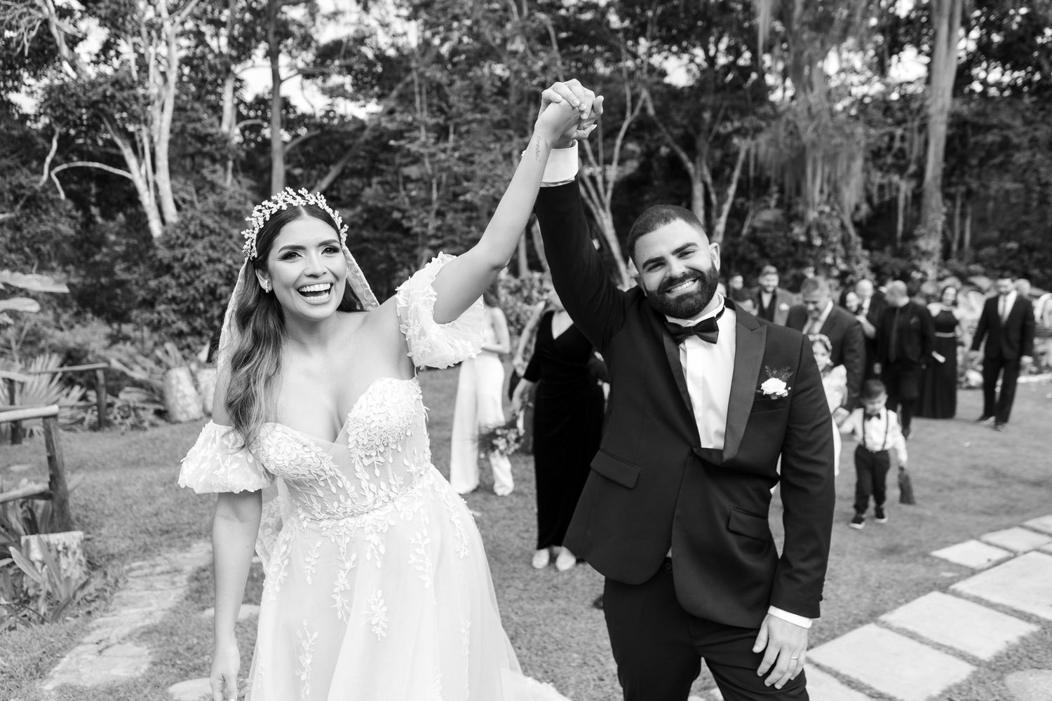 A joyful wedding party walks together through a garden setting in black and white formal attire.