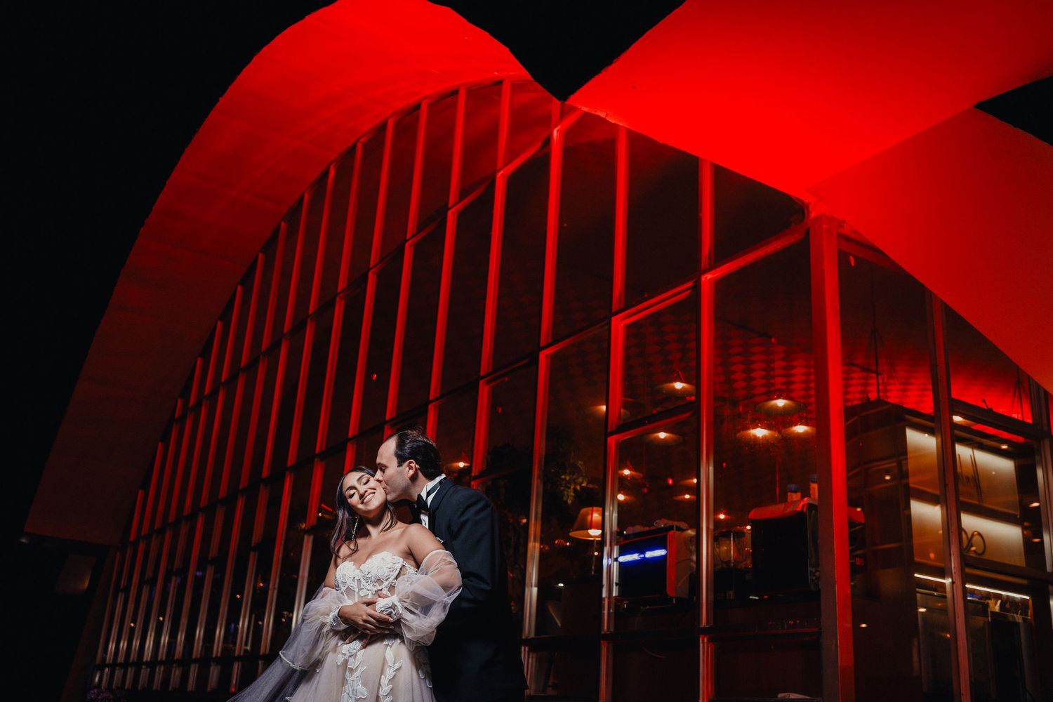 A couple embraces in front of a modern building illuminated with dramatic red lighting at night.