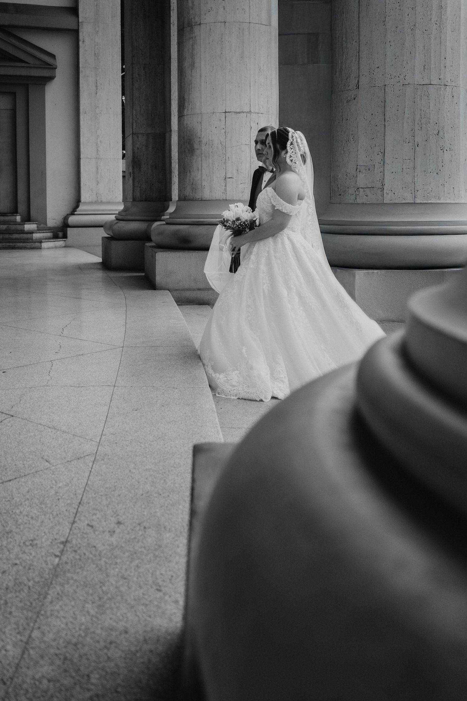 A bride in a flowing white wedding dress and veil stands elegantly between classical stone columns.