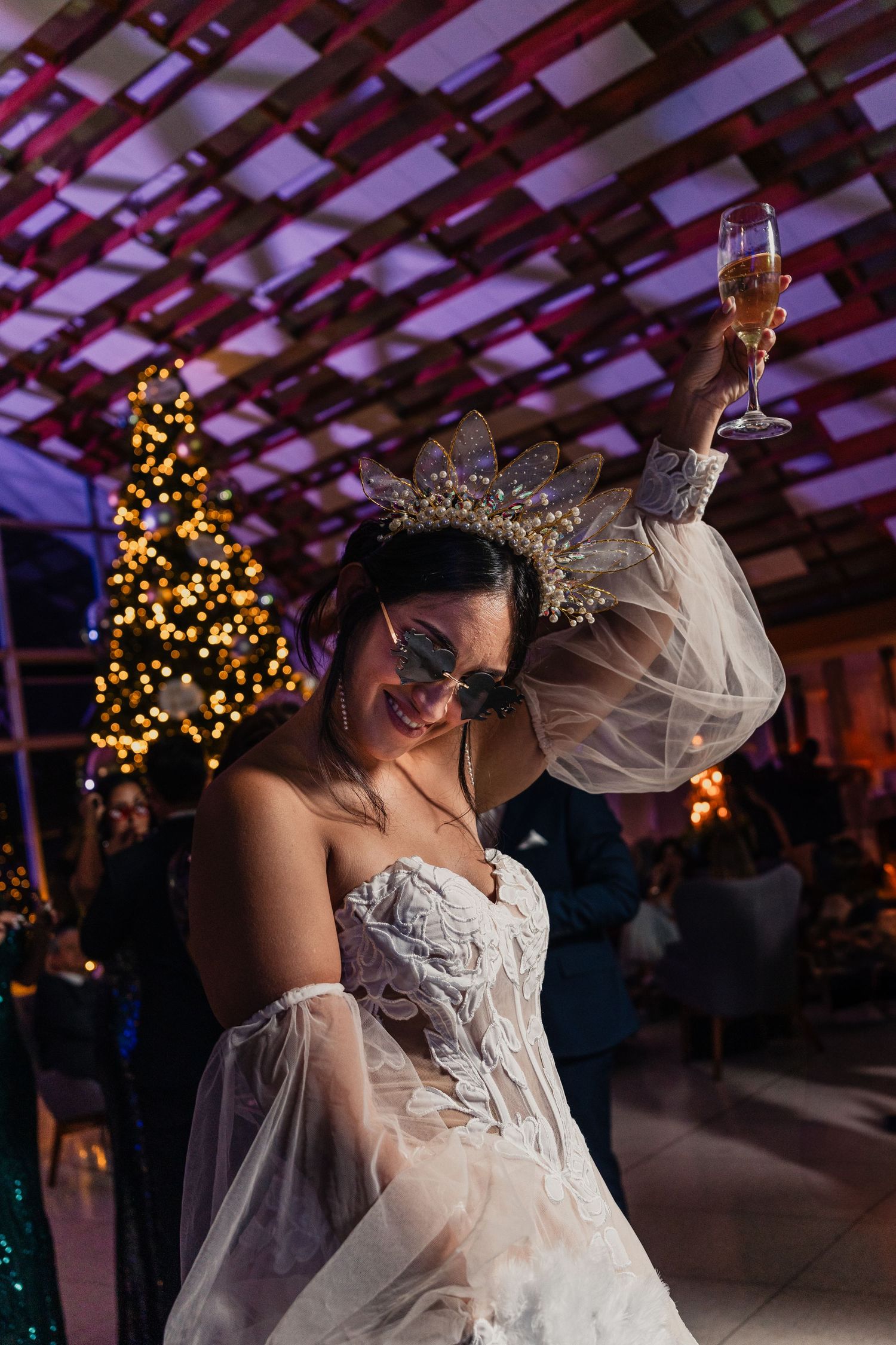 A bride in a white gown raises a champagne glass while dancing at a reception with a Christmas tree in the background.